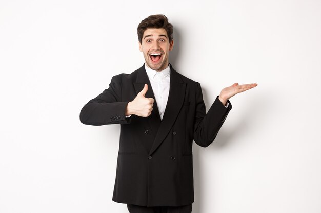 Portrait of hadnsome bearded man in formal suit, showing thumb-up and holding product in hand over white copy space, recommending product, standing over white background.