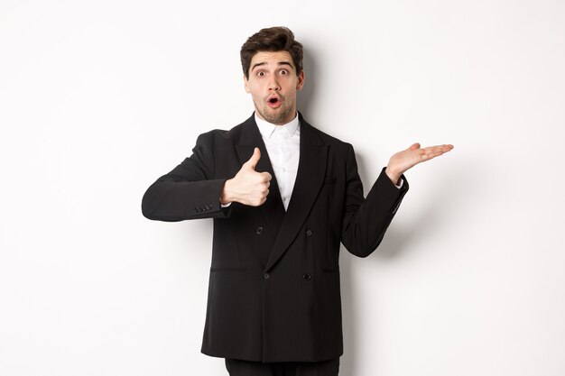 Portrait of hadnsome bearded man in formal suit, showing thumb-up and holding product in hand over white copy space, recommending product, standing over white background