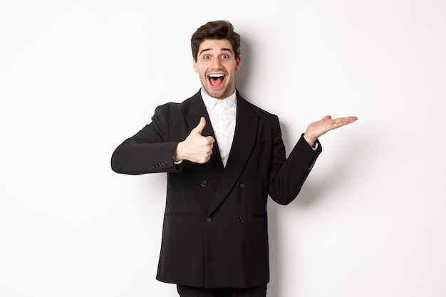 Portrait of hadnsome bearded man in formal suit, showing thumb-up and holding product in hand over white copy space, recommending product, standing over white background