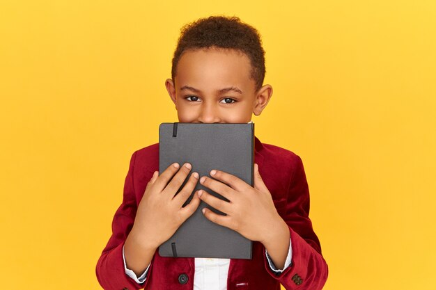 Portrait of handsome Afro American boy having playful eyes covering face with black copy book. black pupil posing isolated holding diary, keeping secret