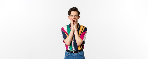 Portrait of handsome androgynous man looking shocked gasping and holding hands on face stare at camera amazed standing over white background