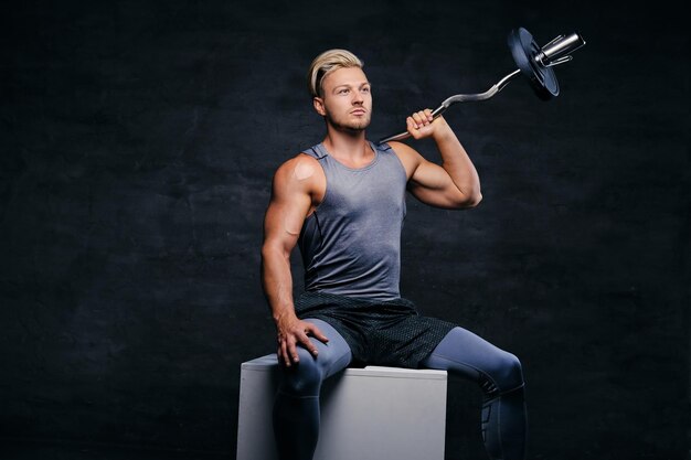 Portrait of a handsome, athletic blond male sits on a white box and holds barbell on his shoulder.