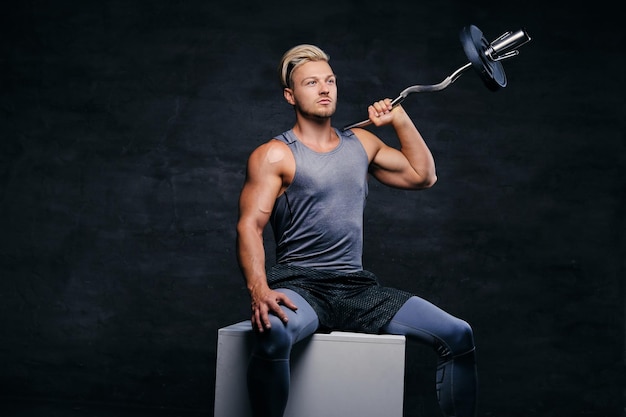 Portrait of a handsome, athletic blond male sits on a white box and holds barbell on his shoulder.