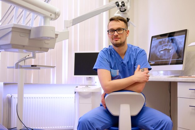 Portrait of a handsome bearded dentist male in glasses wearing a blue uniform, sitting in a dentist office. Looking at camera.