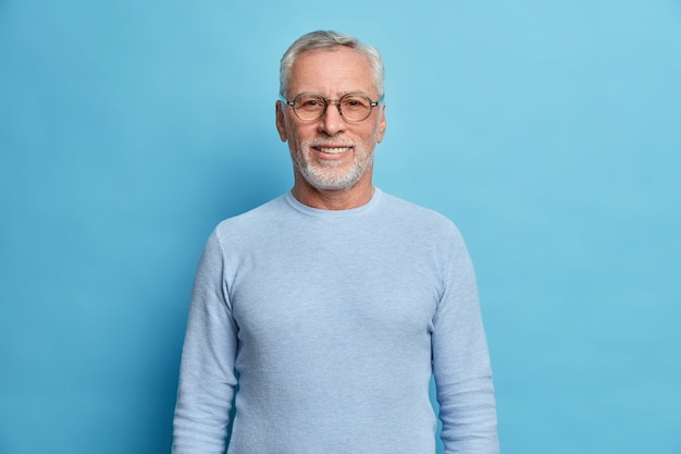 Portrait of handsome bearded european man with grey hair and beard smiles pleasantly looks directly at front being in good mood has lucky day wears spectacles and sweater isolated over blue wall