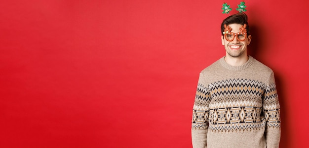 Free photo portrait of handsome bearded guy in party glasses and winter sweater, smiling pleased, celebrating new year, wishing merry christmas, standing over red background