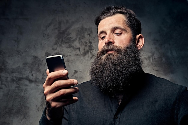 Portrait of a handsome bearded hipster dressed in a black shirt, using a smartphone, standing in a studio. Isolated on a dark background.