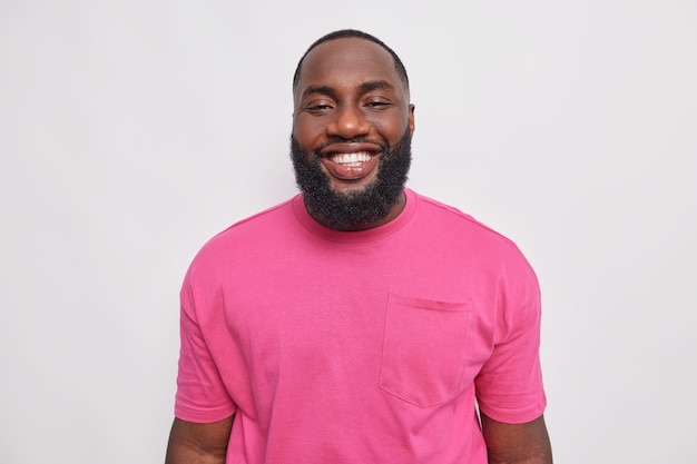Портрет красивого бородатого мужчины, радостно улыбающегося впереди, показывает, что белые идеальные зубы имеют хорошее настроение, чувствует себя довольным, одетый в простую розовую футболку, позирует в помещении