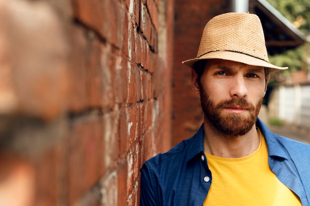 Foto gratuita ritratto di bell'uomo barbuto con cappello di paglia accanto al muro di mattoni