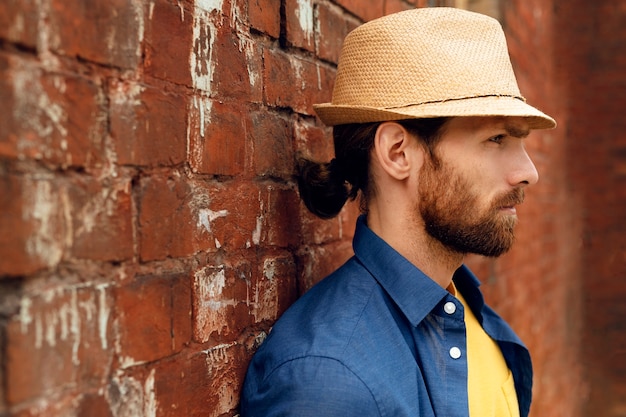 Foto gratuita ritratto di bell'uomo barbuto con cappello di paglia accanto al muro di mattoni