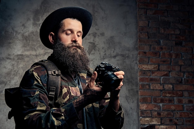 Портрет красивого бородатого путешественника в шляпе и камуфляжной куртке, с рюкзаком и татуированными руками, держит фотоаппарат. Изолированные на темном фоне.
