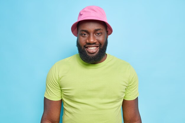 Portrait of handsome black man smiles happily has thick beard big cheerful smile on facewears panama and green t shirt smiles toothily enjoys good day isolated over blue wall