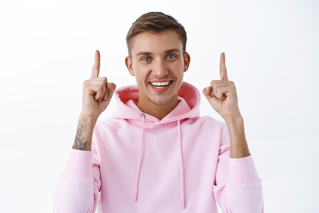 Portrait of handsome blond young caucasian guy, pink hoodie, blue eyes, smiling broadly, pointing fingers up, showing way to promo on top, company banner, click link, standing white wall