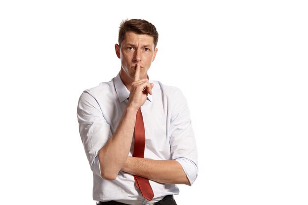 Portrait of a handsome brunet fellow with brown eyes, wearing in a white shirt and a red tie. He is showing a quiet sign while posing in a studio isolated over a white background. Concept of gesticula