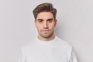 Portrait of handsome brunet unshaven adult man looks with calm confident expression has serious look wears casual jumper poses for making photo against white background being hard to impress