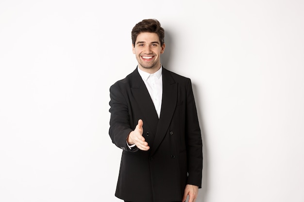Portrait of handsome businessman in black suit, extending hand for handshake, greet business partners and smiling, welcome to company, standing over white background.