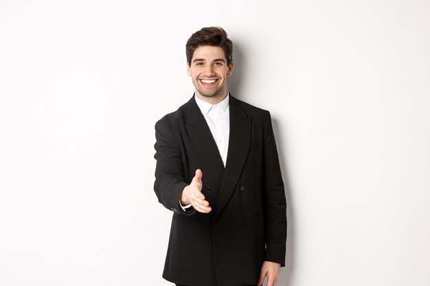 Портрет красивого бизнесмена в черном костюме, протягивающего руку для рукопожатия, приветствуя деловых партнеров и улыбаясь, добро пожаловать в компанию, стоя на белом фоне