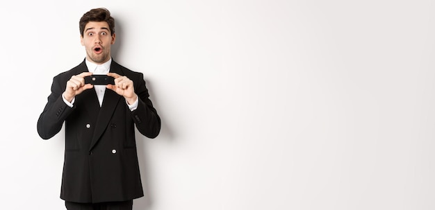 Free photo portrait of handsome businessman in black suit showing credit card and looking amazed standing over