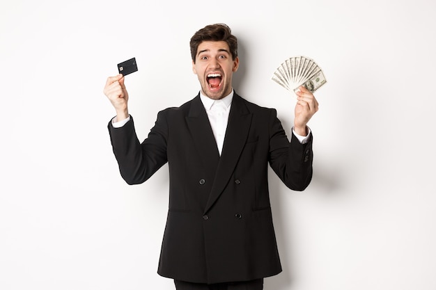 Portrait of handsome businessman in black suit, showing credit card and money, shouting for joy and excitement, standing against white background.