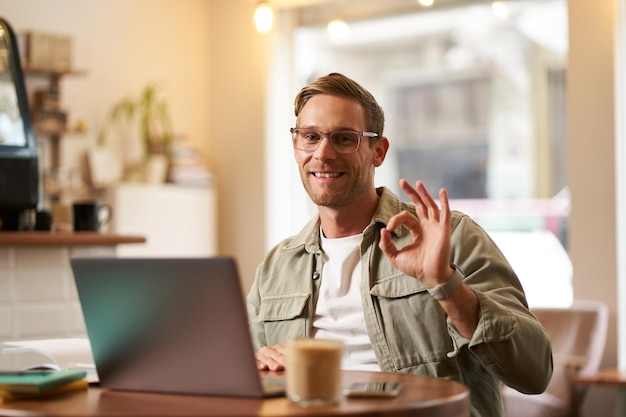 Портрет симпатичного бизнесмена в очках, показывающего знак "окей", сидящего с ноутбуком в кафе