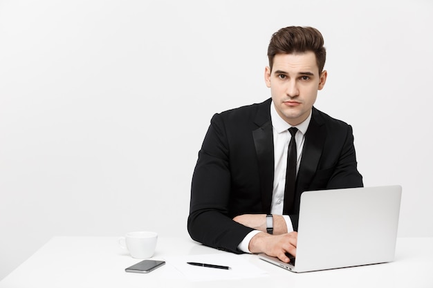 彼は彼の机でコンピューターで作業しながらスマートフォンを持っているハンサムなビジネスマンの肖像画です...