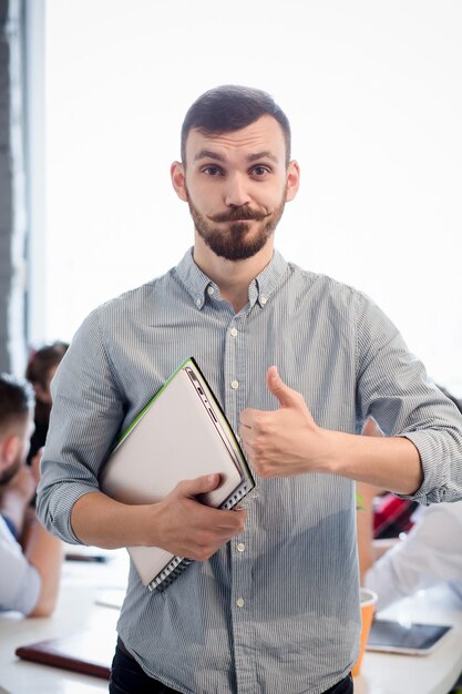 Portrait of handsome businessman showing thumb-up while holding clipboard in office interior.