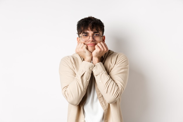 Portrait of handsome caucasian man watching something lovely and cute, squeeze cheeks and lean on hands, looking with admiration and interest at camera, standing on white background.