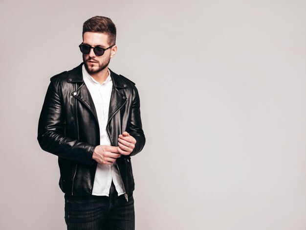 Portrait of handsome confident model Sexy stylish man dressed in biker leather jacket and black jeans Fashion hipster male isolated on grey background in studio in sunglasses Isolated