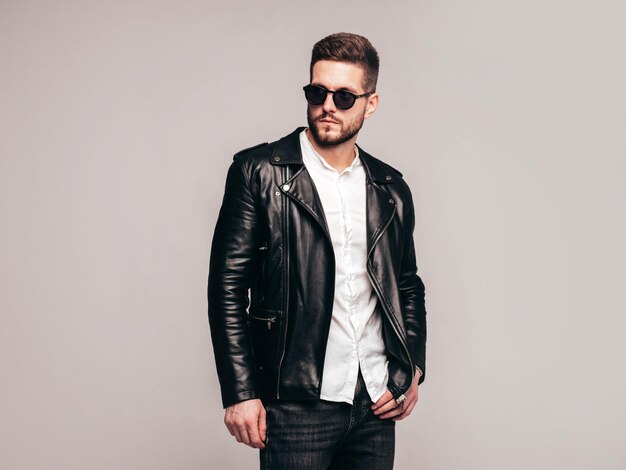 Portrait of handsome confident model Sexy stylish man dressed in biker leather jacket and black jeans Fashion hipster male isolated on grey background in studio in sunglasses Isolated