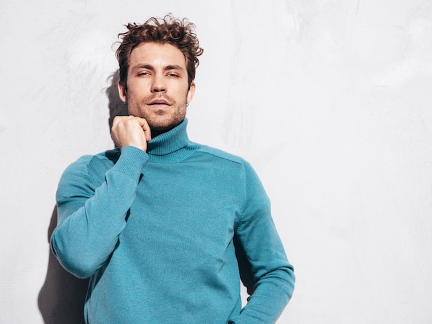 Portrait of handsome confident model Sexy stylish man dressed in blue sweater and jeans Fashion hipster male with curly hairstyle posing near grey wall in studio Isolated