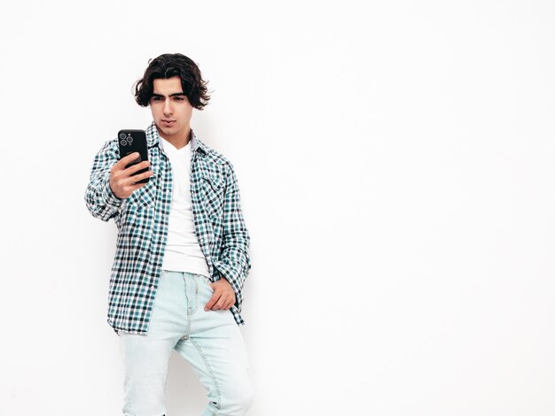 Portrait of handsome confident model Sexy stylish man dressed in checkered shirt and jeans Fashion hipster male posing near white wall in studio Taking selfie photos