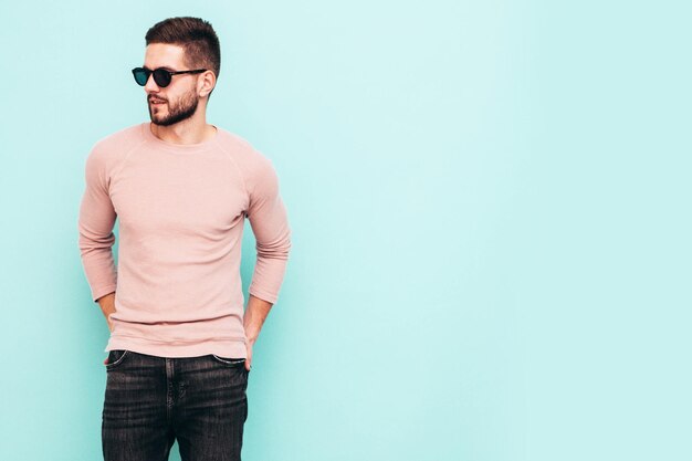Portrait of handsome confident model Sexy stylish man dressed in pink sweater and jeans Fashion hipster male posing near blue wall in studio In sunglasses Isolated