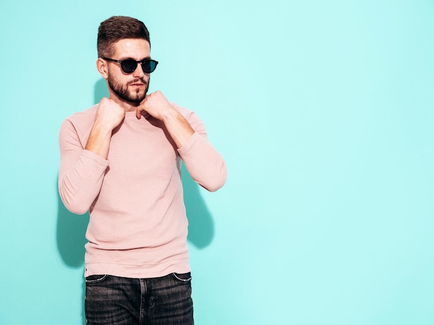 잘 생긴 자신감 모델의 초상화 핑크 스웨터와 청바지를 입고 섹시 한 세련 된 남자 패션 힙 스터 남성 스튜디오에서 파란색 벽 근처 포즈 선글라스 절연