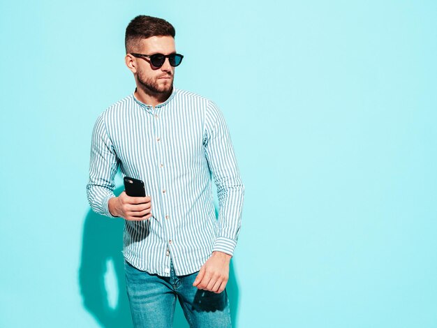 Portrait of handsome confident model Sexy stylish man dressed in shirt and jeans Fashion hipster male posing near blue wall in studio in sunglasses Holding smartphone