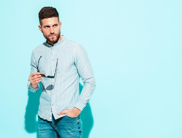 Portrait of handsome confident model Sexy stylish man dressed in shirt and jeans Fashion hipster male posing near blue wall in studio