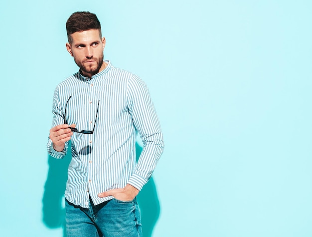 잘 생긴 자신감 모델의 초상화 셔츠와 청바지를 입고 섹시 한 세련 된 남자 패션 힙 스터 남성 스튜디오에서 파란색 벽 근처 포즈