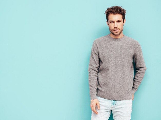 Portrait of handsome confident model Sexy stylish man dressed in sweater and jeans Fashion hipster male with curly hairstyle posing near blue wall in studio Isolated