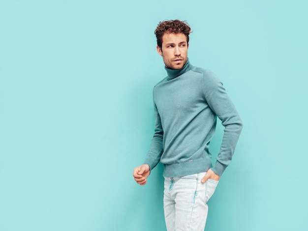 Ritratto di bel modello fiducioso uomo alla moda sexy vestito in maglione e jeans moda hipster maschio con acconciatura riccia in posa vicino al muro blu in studio isolato
