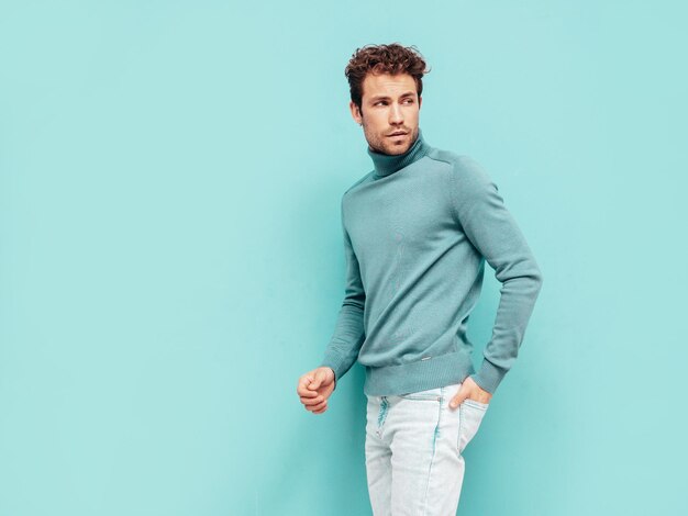 잘 생긴 자신감 모델의 초상화 스웨터와 청바지를 입고 섹시 한 세련 된 남자 패션 힙 스터 남성 곱슬 헤어스타일 절연 스튜디오에서 파란색 벽 근처 포즈