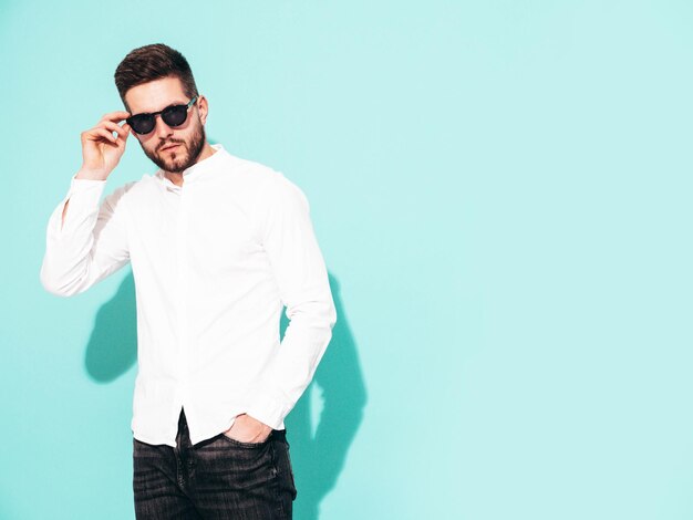 잘 생긴 자신감 모델의 초상화 흰색 셔츠와 청바지를 입고 섹시 한 세련 된 남자 패션 힙 스터 남성 선글라스에 절연 스튜디오에서 파란색 벽 근처 포즈