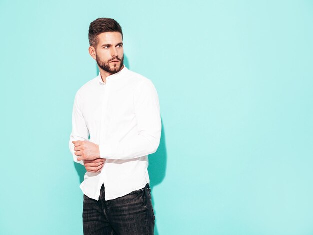 잘 생긴 자신감 모델의 초상화 흰색 셔츠와 청바지를 입고 섹시 한 세련 된 남자 패션 힙 스터 남성 스튜디오 격리에 파란색 벽 근처 포즈
