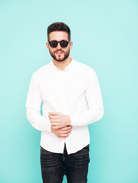 Portrait of handsome confident model Sexy stylish man dressed in white shirt and jeans Fashion hipster male posing near blue wall in studio in sunglasses Isolated