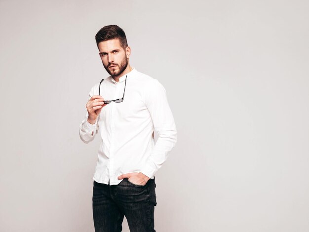 Portrait of handsome confident model Sexy stylish man dressed in white shirt and jeans Fashion hipster male posing in studio on grey background Holding sunglasses Isolated