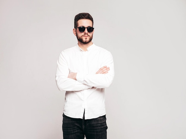 Portrait of handsome confident model Sexy stylish man dressed in white shirt and jeans Fashion hipster male posing in studio on grey background In sunglasses Isolated