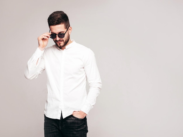 Portrait of handsome confident model Sexy stylish man dressed in white shirt and jeans Fashion hipster male posing in studio on grey background In sunglasses Isolated