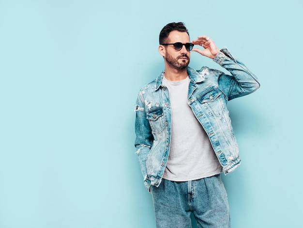 Портрет красивой уверенной в себе стильной хипстерской модели ламберсексуала Сексуальный мужчина, одетый в джинсовую куртку Модный мужчина, изолированный на синей стене в студии В солнцезащитных очках