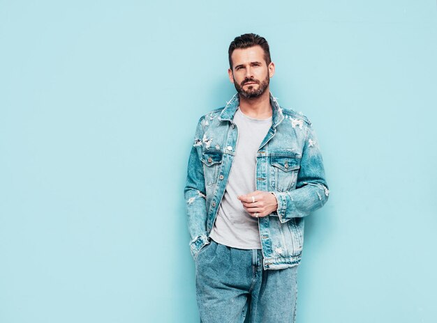 Portrait of handsome confident stylish hipster lambersexual model Sexy man dressed in jeans jacket Fashion male isolated on blue wall in studio