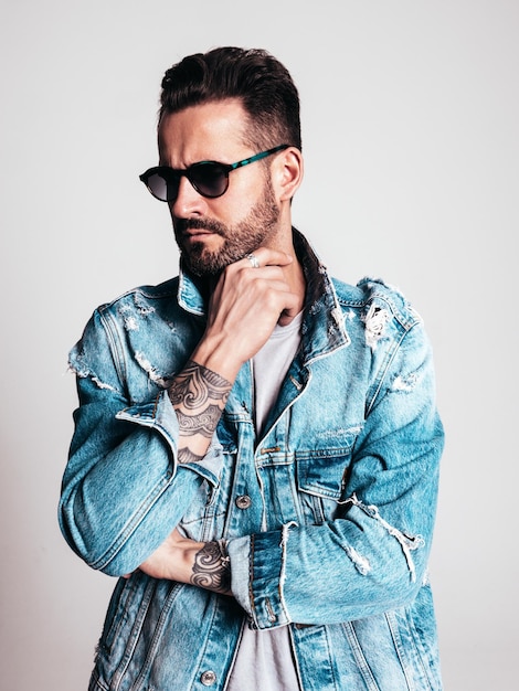 Portrait of handsome confident stylish hipster lambersexual model Sexy man dressed in jeans jacket Fashion male isolated on grey background in studio in sunglasses