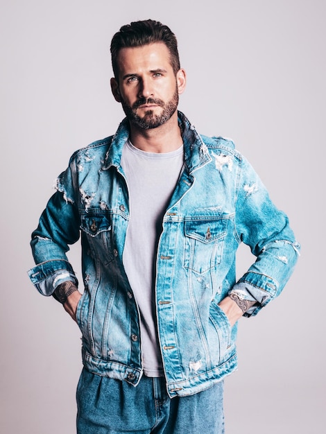 Portrait of handsome confident stylish hipster lambersexual model Sexy man dressed in jeans jacket Fashion male isolated on grey background in studio