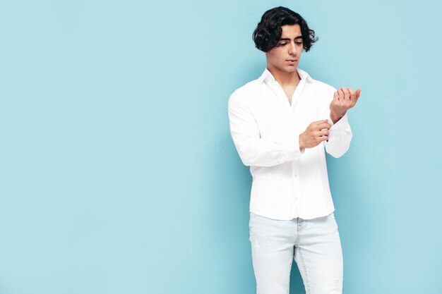 Portrait of handsome confident stylish hipster lambersexual model Sexy man dressed in summer white shirt and jeans clothes Fashion male isolated in studio Posing near blue wall